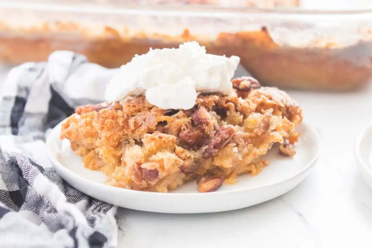A slice of Pumpkin Crunch Cake on a white plate, topped with whipped cream, with a checkered cloth beside it and a baking dish in the background.
