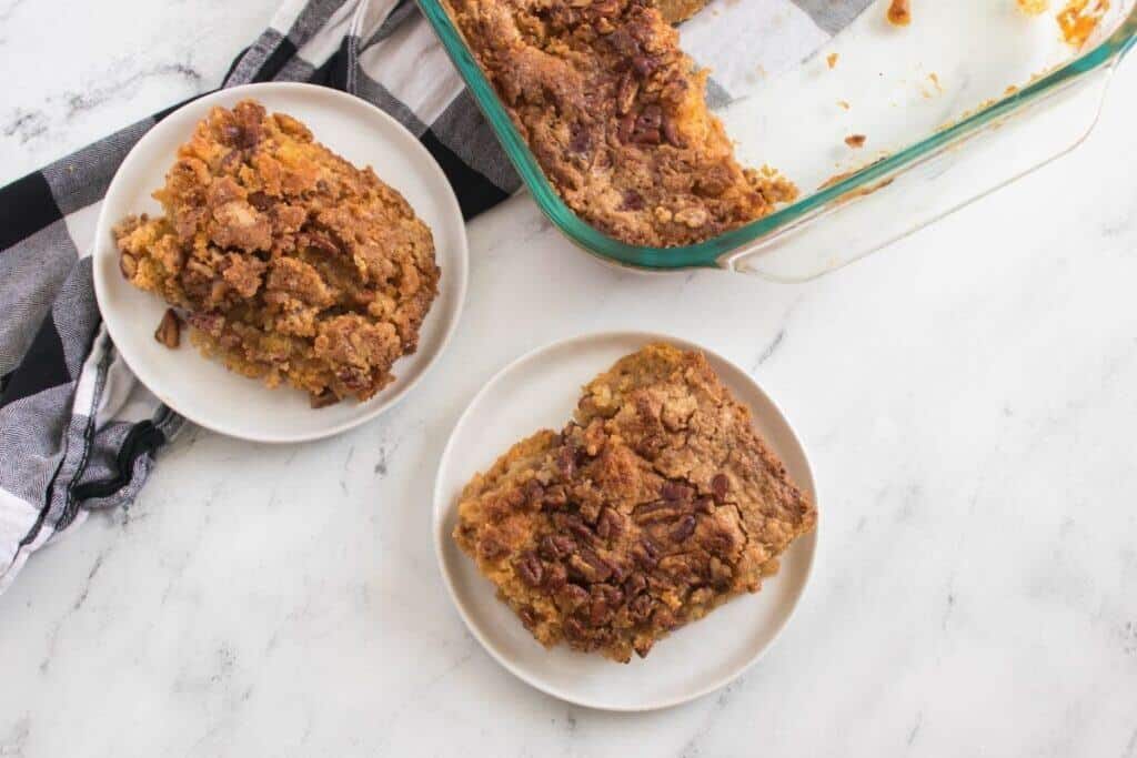 Two plates with servings of Pumpkin Crunch Cake next to a glass baking dish, all placed on a white marble surface with a black and white cloth nearby.