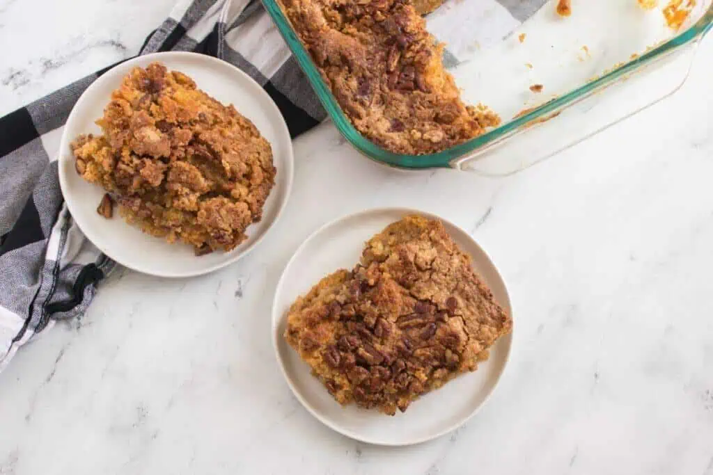 Two plates with servings of Pumpkin Crunch Cake next to a glass baking dish, all placed on a white marble surface with a black and white cloth nearby.
