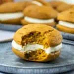 Pumpkin whoopie pie with cream filling on a metal plate, partially eaten to reveal its soft, center. More pies are blurred in the background.