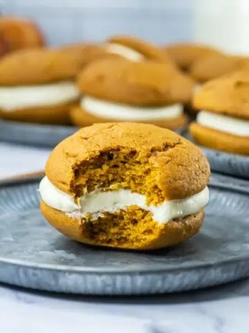 Pumpkin whoopie pie with cream filling on a metal plate, partially eaten to reveal its soft, center. More pies are blurred in the background.