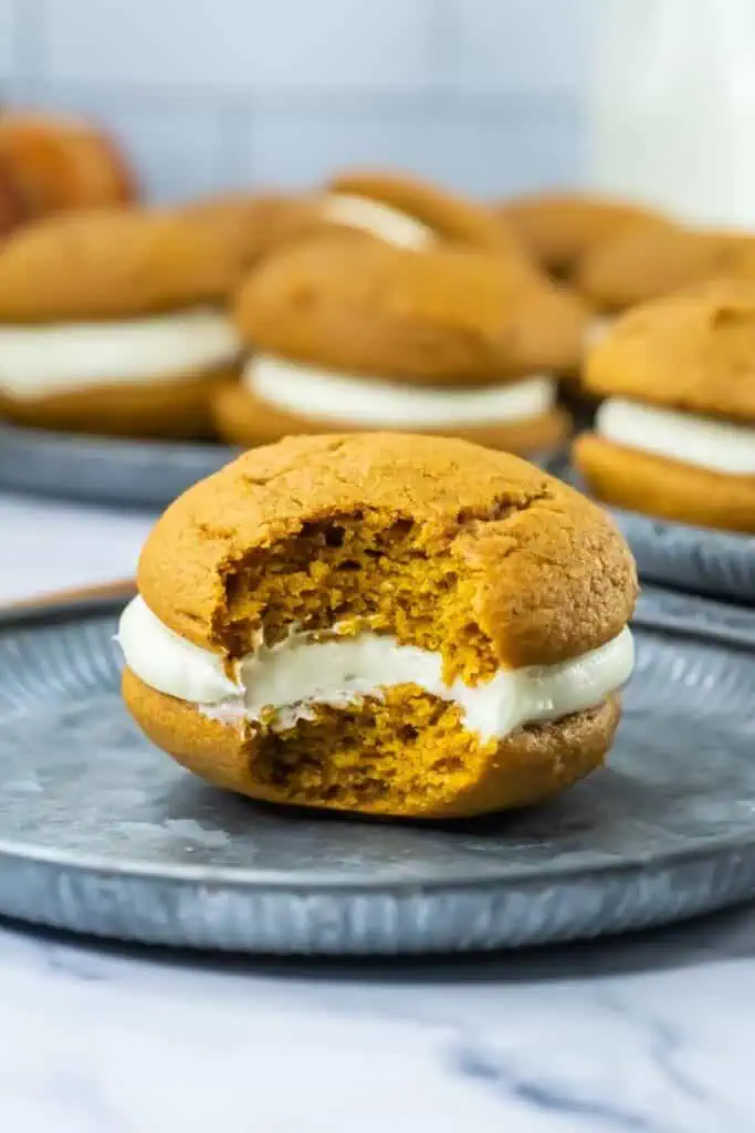 Pumpkin whoopie pie with cream filling on a metal plate, partially eaten to reveal its soft, center. More pies are blurred in the background.