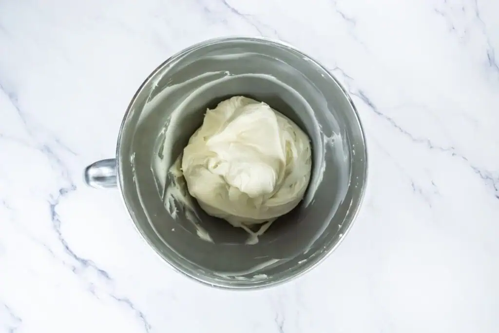 Bowl of smooth cream cheese frosting in a metal mixing bowl on a marble surface.