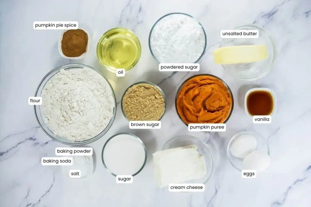 Overhead view of labeled pumpkin whoopie pie ingredients in glass bowls on a marble surface.