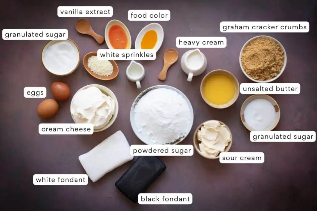 Assorted baking ingredients, including sugars, eggs, cream cheese, heavy cream, sour cream, butter, food color, sprinkles, vanilla extract, graham cracker crumbs, and white and black fondant.