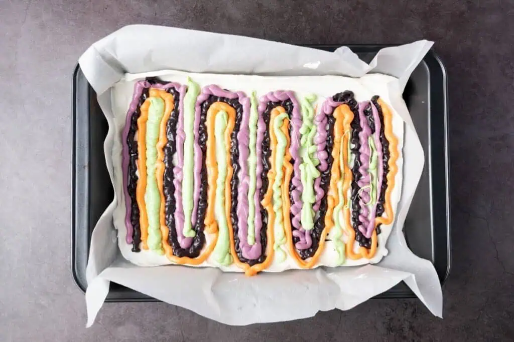 A rectangular baking pan lined with parchment paper holds a dessert topped with wavy lines of orange, purple, green, and black glaze.
