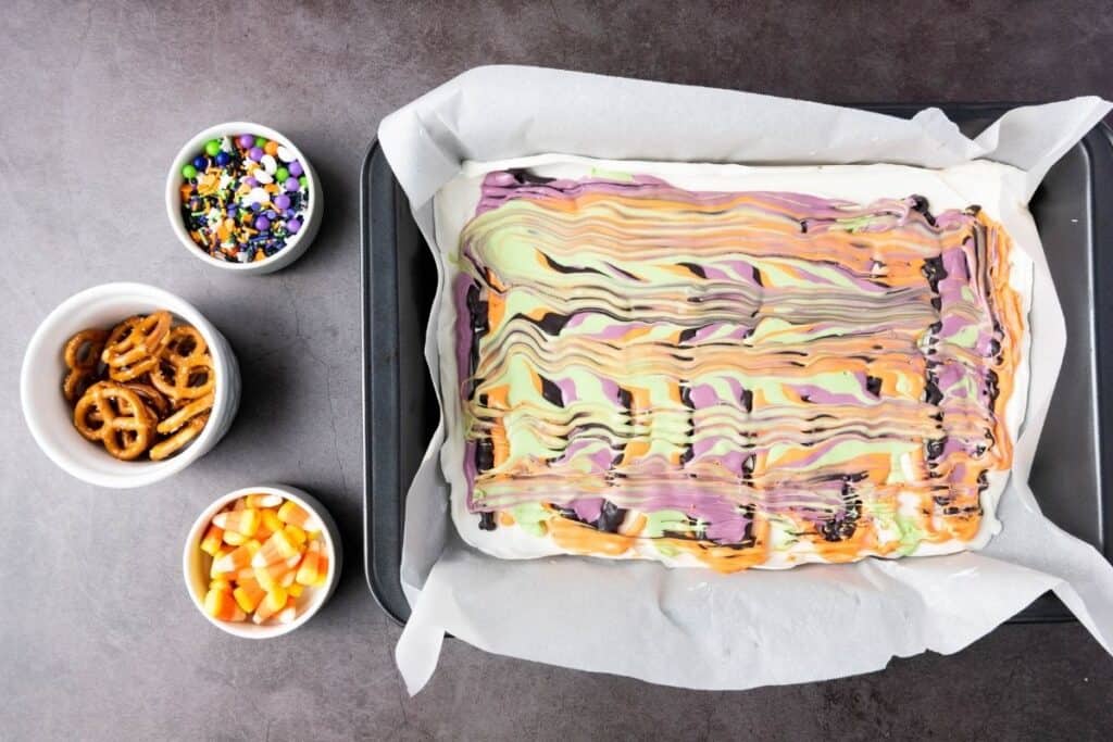 A baking tray with swirled Halloween-colored bark on parchment paper, next to small bowls of Halloween sprinkles, pretzels, and candy corn.