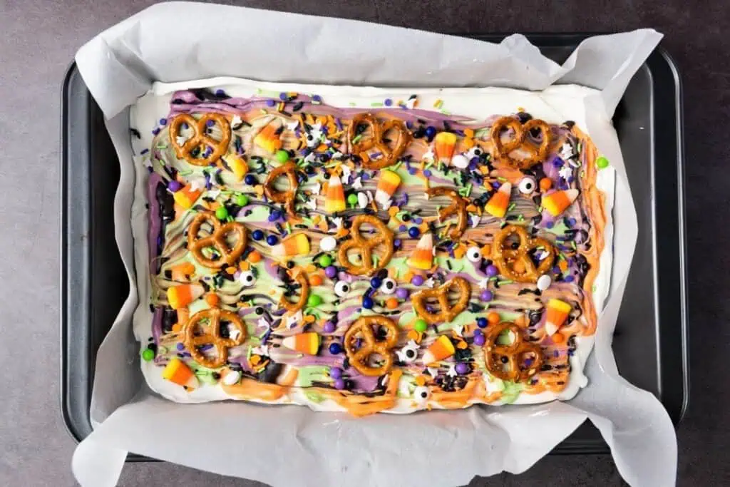 A tray of Halloween-themed dessert bark topped with pretzels, candy corn, colorful drizzles, and festive sprinkles on parchment paper.