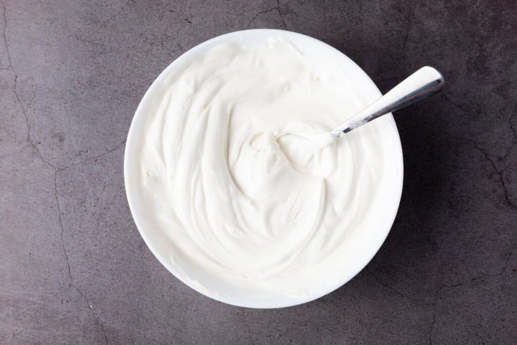 A white bowl filled with melted white candy with a metal spoon resting inside, placed on a gray textured surface.