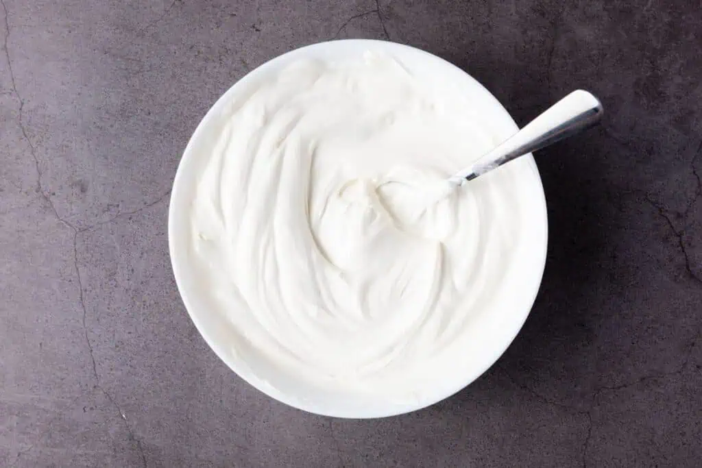 A white bowl filled with melted white candy with a metal spoon resting inside, placed on a gray textured surface.