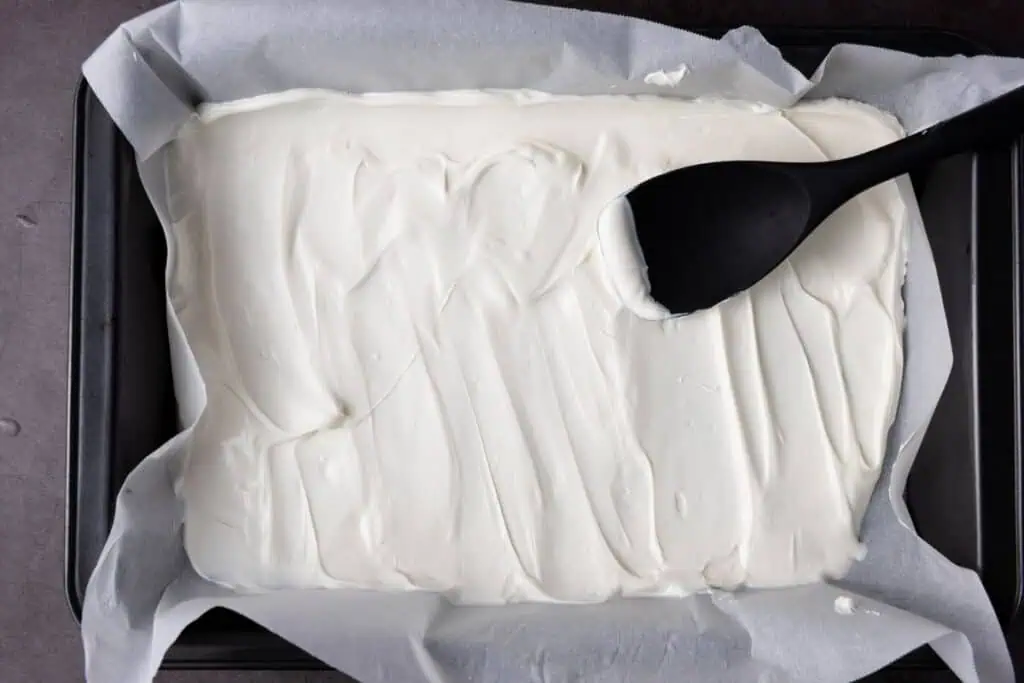 A rectangular baking pan lined with parchment paper contains a smooth layer of white melted candy, being spread with a black spatula.
