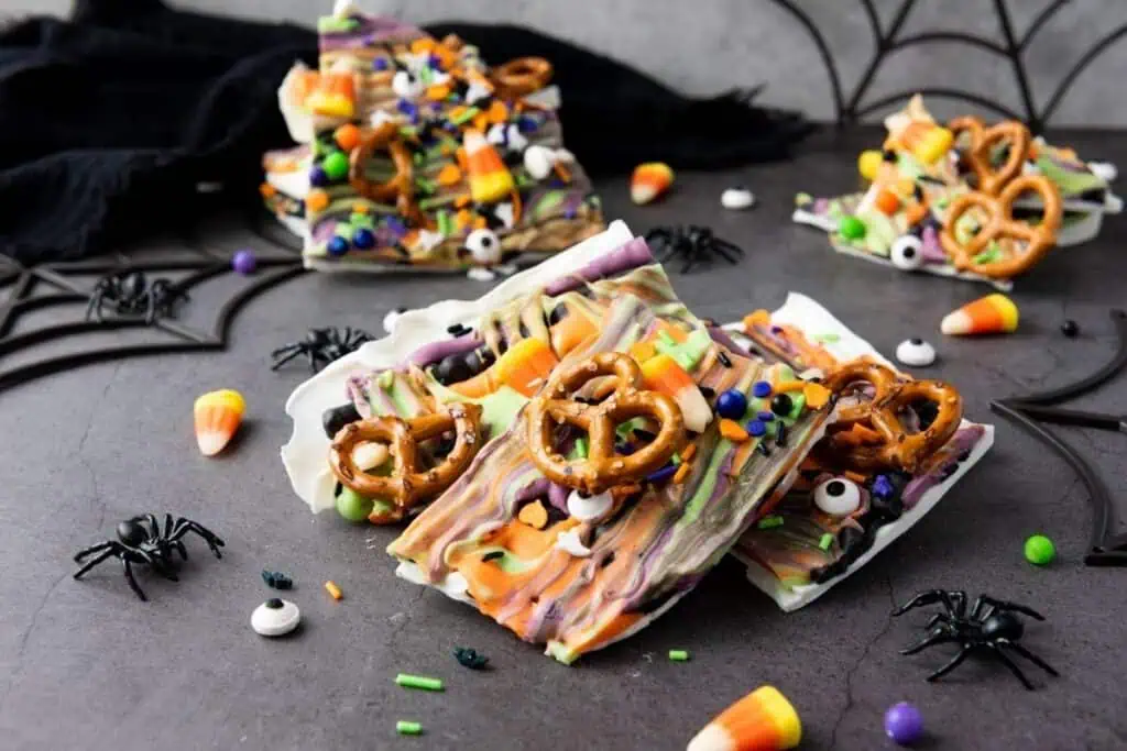 Colorful Halloween-themed bark topped with pretzels, candy eyes, candy corn, and sprinkles, displayed on a dark surface with plastic spiders and web decorations.