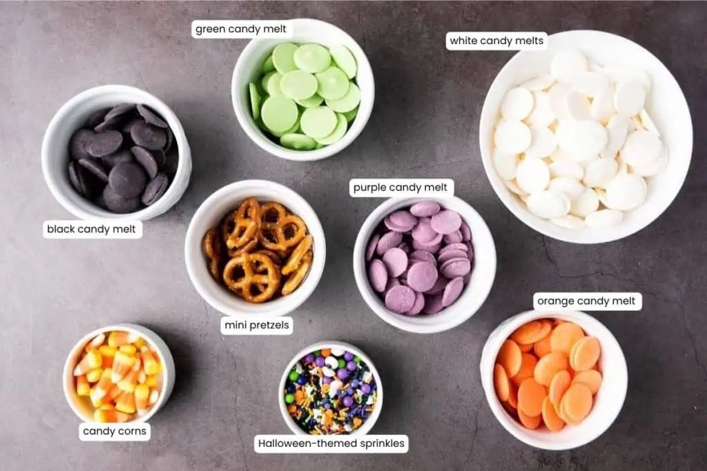 Eight bowls containing black, green, white, purple, and orange candy melts, mini pretzels, candy corns, and Halloween-themed sprinkles are arranged on a gray surface.