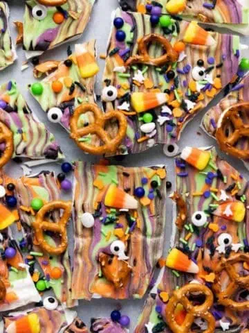 Colorful Halloween bark topped with candy corn, pretzels, candy eyes, sprinkles, and chocolate, broken into pieces on a flat surface.