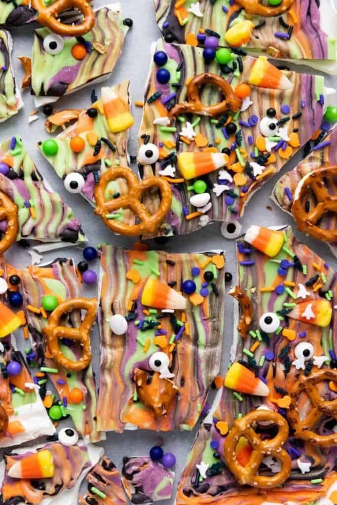 Colorful Halloween bark topped with candy corn, pretzels, candy eyes, sprinkles, and chocolate, broken into pieces on a flat surface.