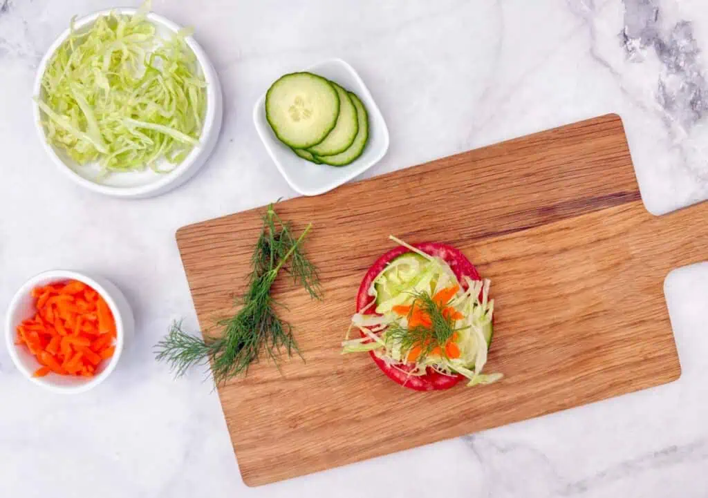 A wooden cutting board with a tomato slice topped with shredded lettuce, cucumber slices, and dill, surrounded by bowls of shredded lettuce, sliced cucumber, and diced orange bell pepper.