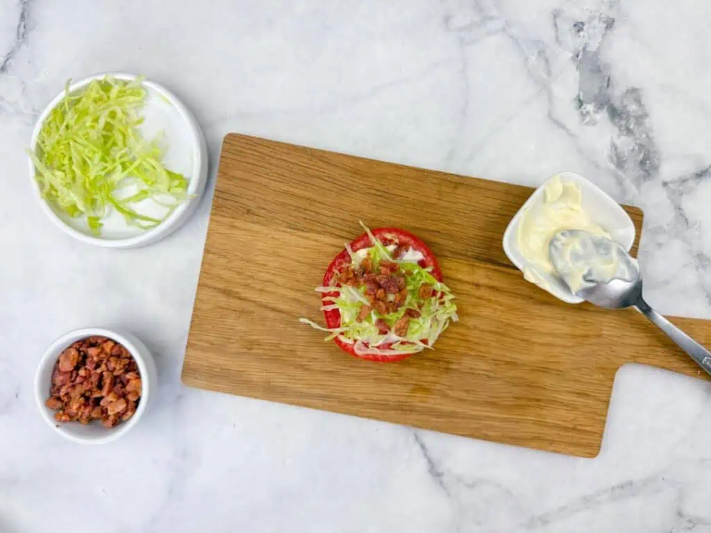 A wooden board with a tomato slice topped with shredded lettuce and bacon bits, next to bowls of lettuce, bacon bits, and a dish of mayonnaise with a spoon on a marble surface.
