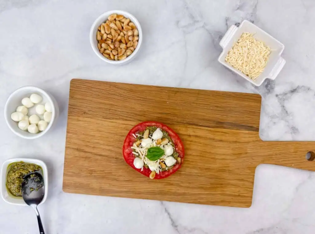 A wooden cutting board holds a tomato slice topped with cheese, mozzarella balls, pine nuts, and basil. Surrounding bowls contain pine nuts, grated cheese, mozzarella balls, and pesto.