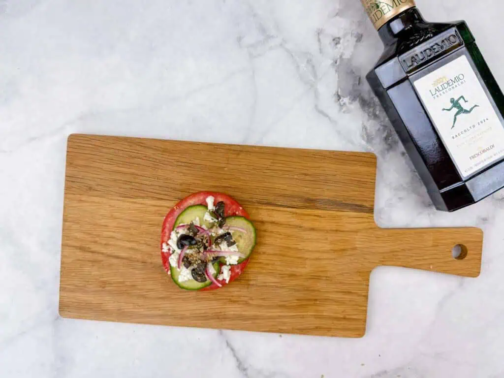 A wooden cutting board with a slice of tomato topped with cucumber, feta, olives, and herbs; a bottle of olive oil is nearby on a marble countertop.