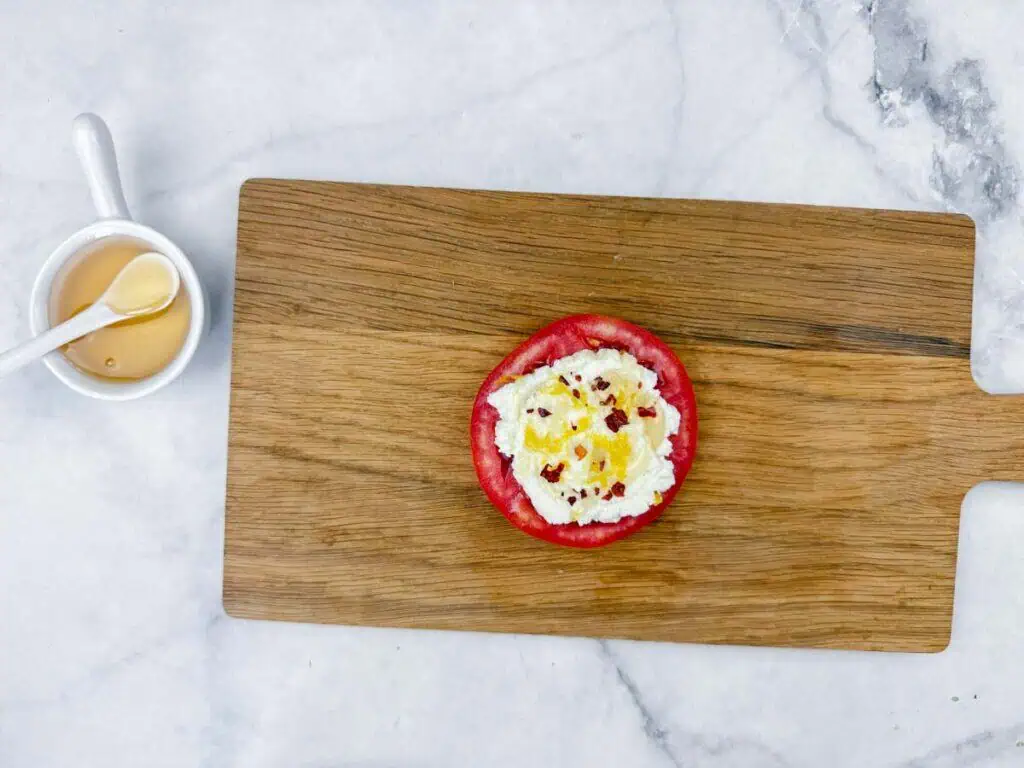 A slice of tomato topped with cottage cheese and seasonings on a wooden cutting board, next to a small cup of honey with a spoon on a marble surface.