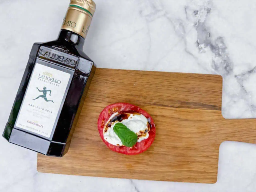 A bottle of Laudemio olive oil sits on a wooden board next to a slice of tomato topped with cheese, basil, and balsamic glaze.