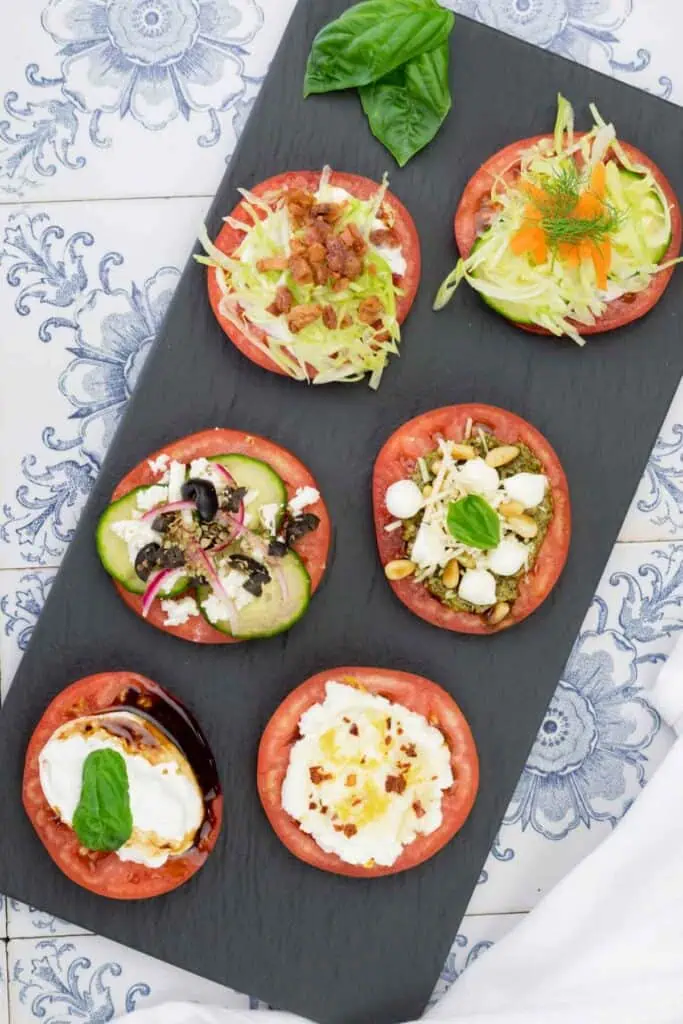 Six tomato slices topped with various fresh ingredients, arranged on a black slate platter over a patterned tile surface.