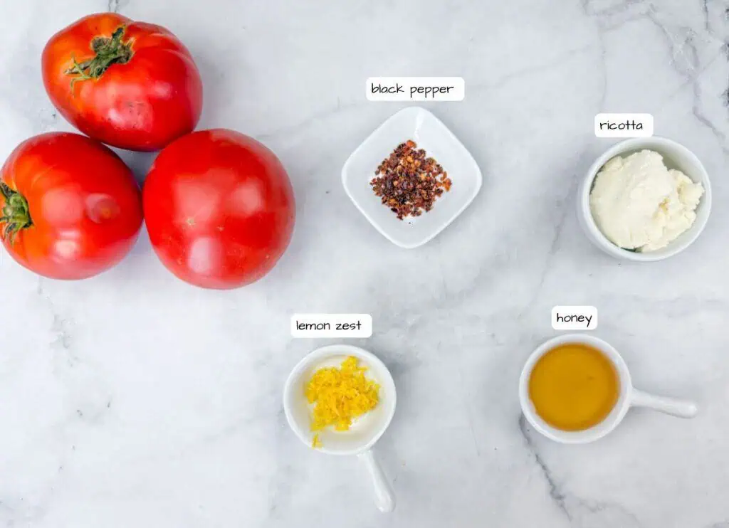 Three tomatoes, a bowl of black pepper, a bowl of ricotta, a bowl of lemon zest, and a bowl of honey are arranged on a marble surface with labeled tags.