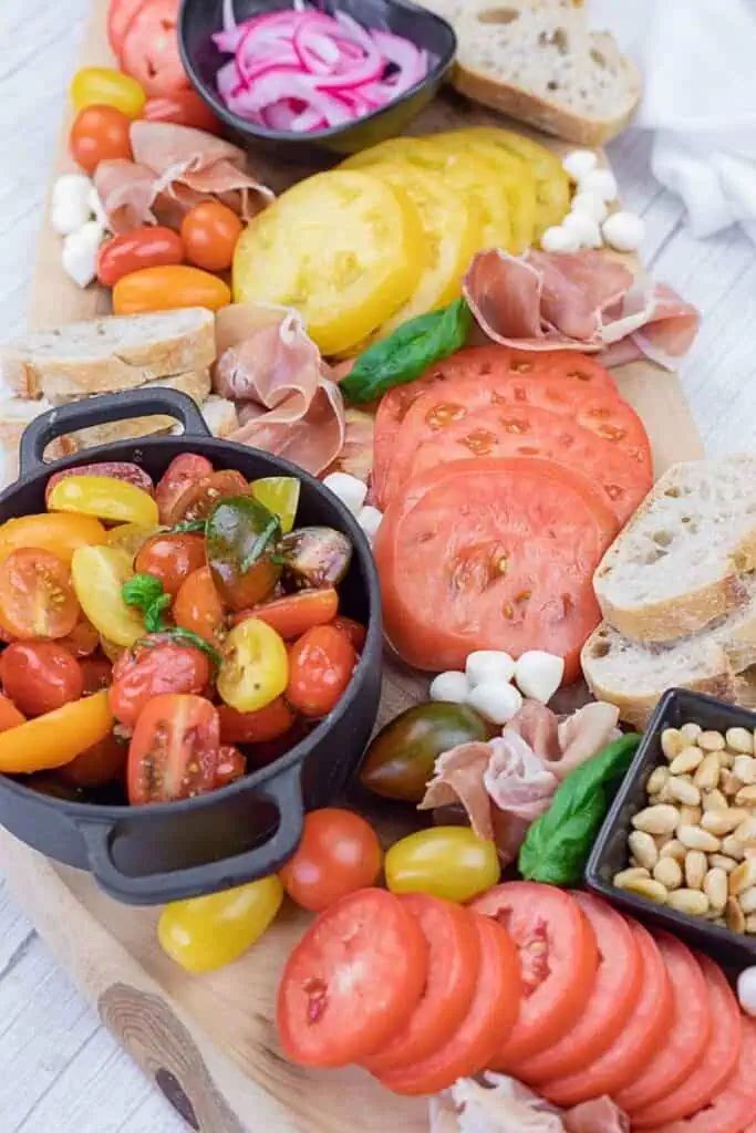 A charcuterie board with sliced tomatoes, cherry tomatoes, mozzarella balls, prosciutto, bread, pickled onions, pine nuts, and basil leaves arranged on a wooden surface.