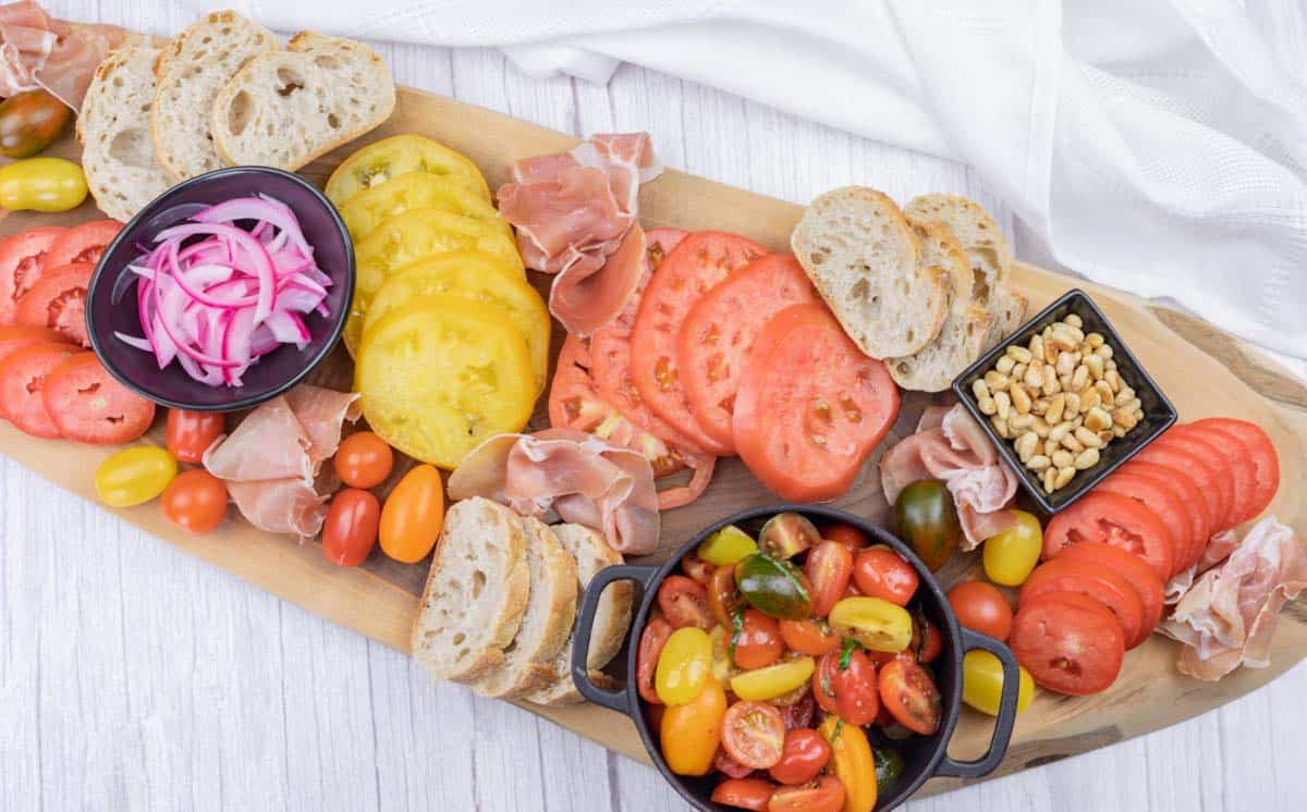 A wooden board with sliced bread, red and yellow tomato slices, cured meat, pickled onions, pine nuts, and a bowl of mixed cherry tomatoes.
