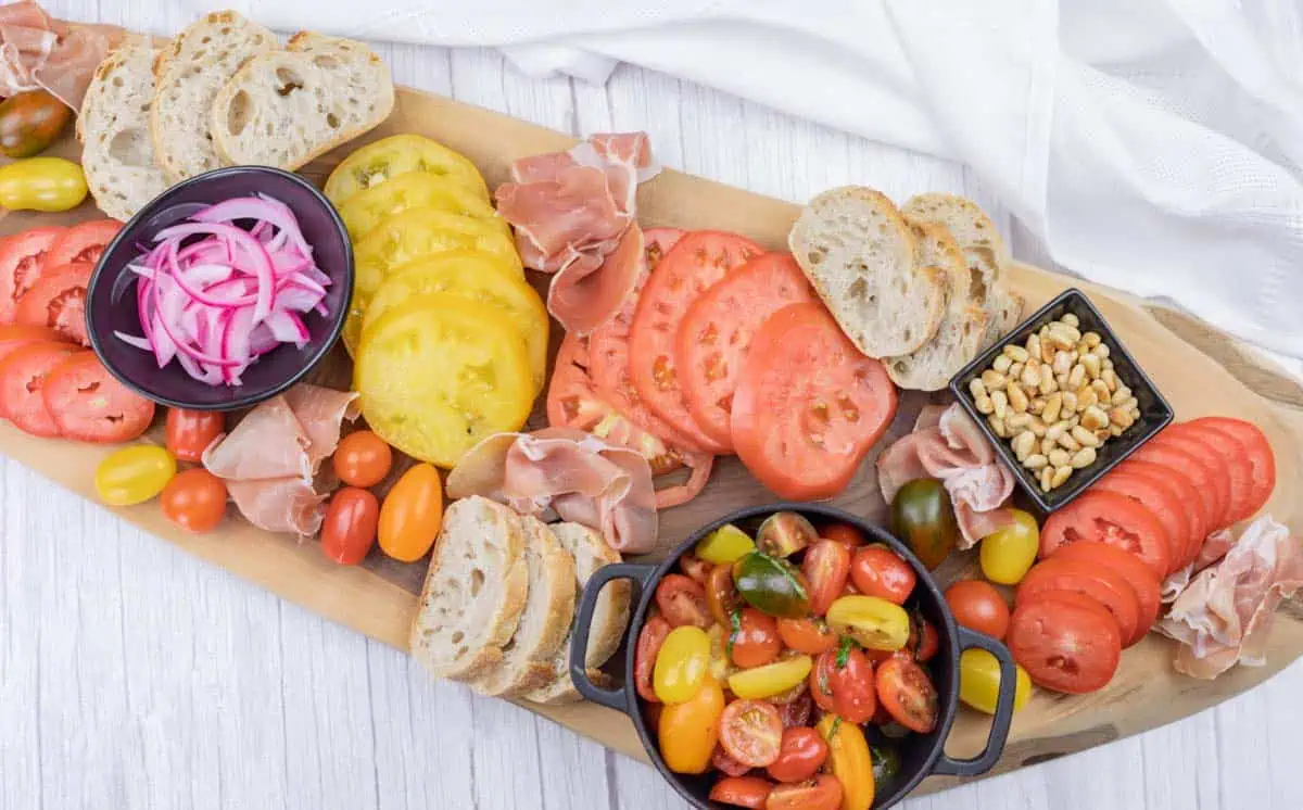 A wooden board with sliced bread, red and yellow tomato slices, cured meat, pickled onions, pine nuts, and a bowl of mixed cherry tomatoes.