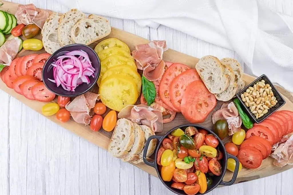 A wooden board with assorted sliced bread, prosciutto, yellow and red tomatoes, pickled onions, cherry tomatoes, pine nuts, and a bowl of tomato salad on a white surface.