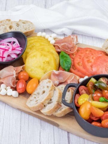 A wooden board displays sliced tomatoes, cherry tomatoes, pickled onions, mozzarella balls, prosciutto, baguette slices, pine nuts, and basil, with napkins in the background.
