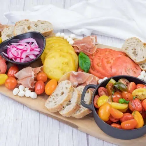 A wooden board displays sliced tomatoes, cherry tomatoes, pickled onions, mozzarella balls, prosciutto, baguette slices, pine nuts, and basil, with napkins in the background.