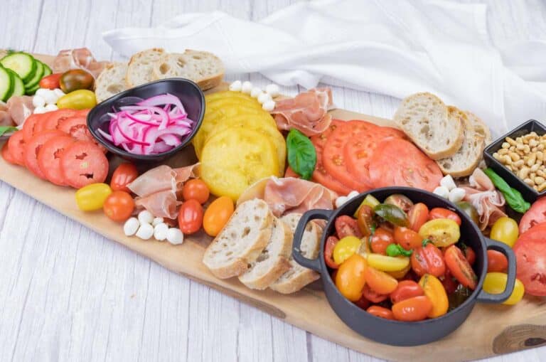 A wooden board displays sliced tomatoes, cherry tomatoes, pickled onions, mozzarella balls, prosciutto, baguette slices, pine nuts, and basil, with napkins in the background.