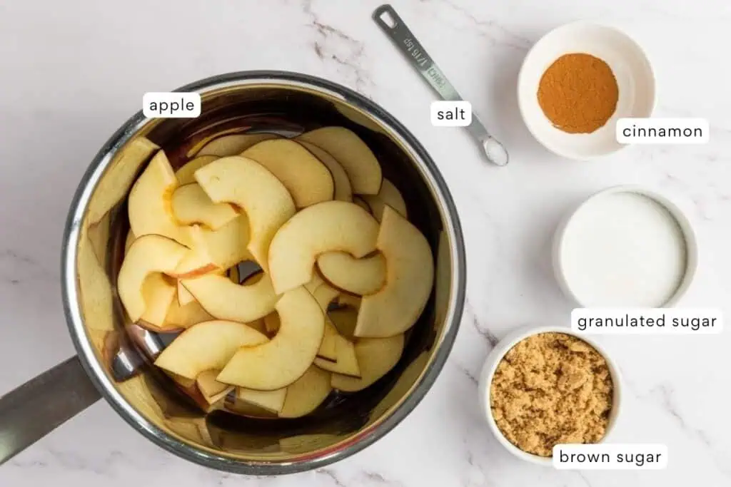 A saucepan with sliced apples next to measured bowls of cinnamon, granulated sugar, brown sugar, and a measuring spoon of salt on a marble surface.