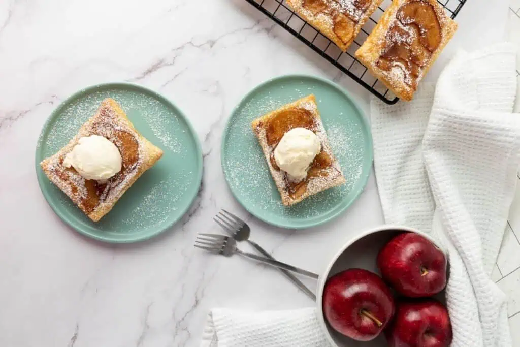 Two plates with apple tarts topped with ice cream, a cooling rack with more tarts, and a bowl of red apples on a white marble surface.