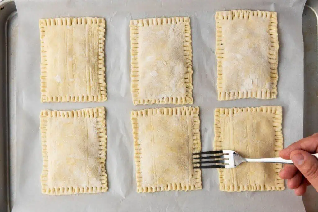 Six rectangular pastry pockets on parchment paper, with a hand using a fork to crimp the edges of one of them.