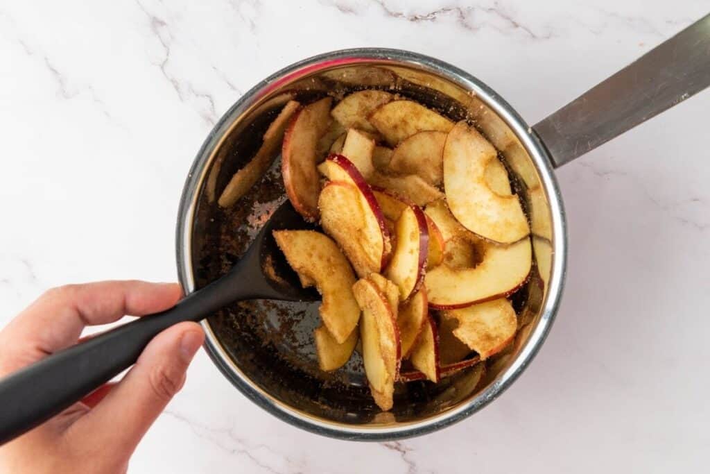 Sliced apples coated with cinnamon and sugar being stirred in a saucepan with a black spoon.