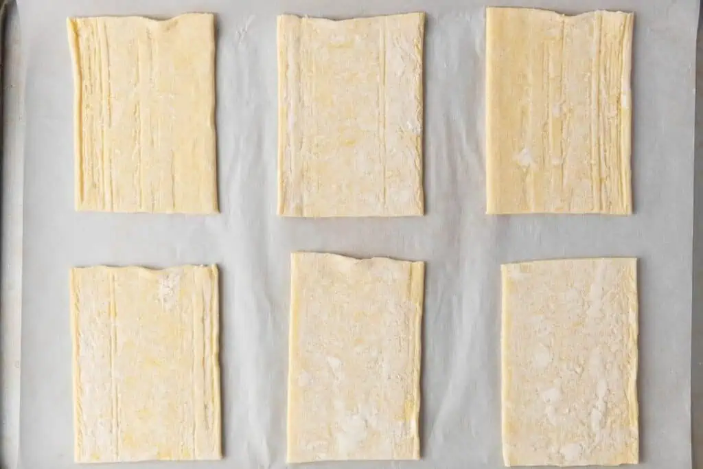 Six rectangular pieces of raw puff pastry dough are arranged in two rows on a parchment-lined baking sheet.
