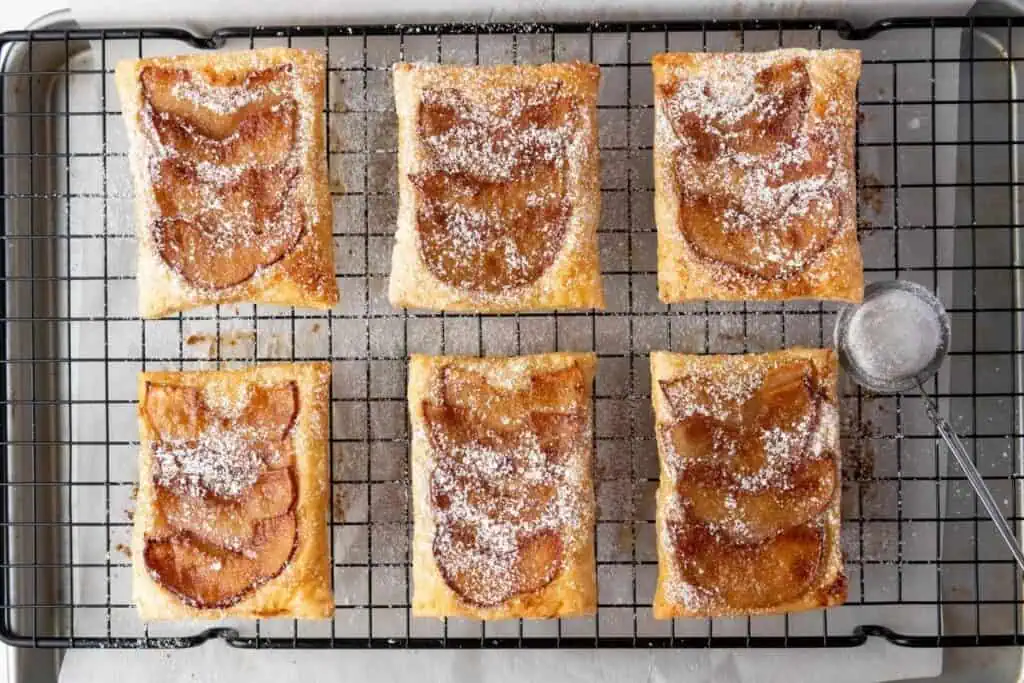 Six rectangular apple pastries topped with powdered sugar are arranged on a metal cooling rack, with a small sifter nearby.