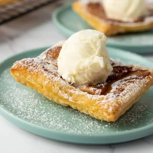 A square piece of puff pastry topped with a scoop of vanilla ice cream and dusted with powdered sugar on a light blue plate.