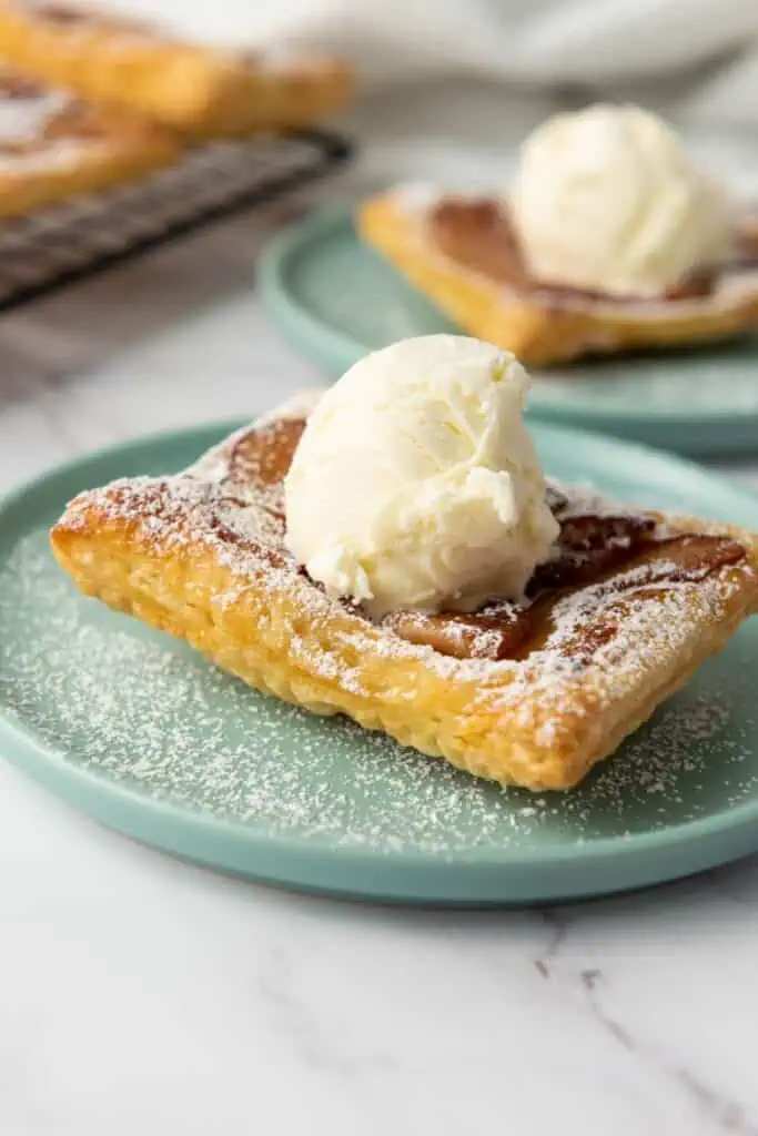 A square piece of puff pastry topped with a scoop of vanilla ice cream and dusted with powdered sugar on a light blue plate.