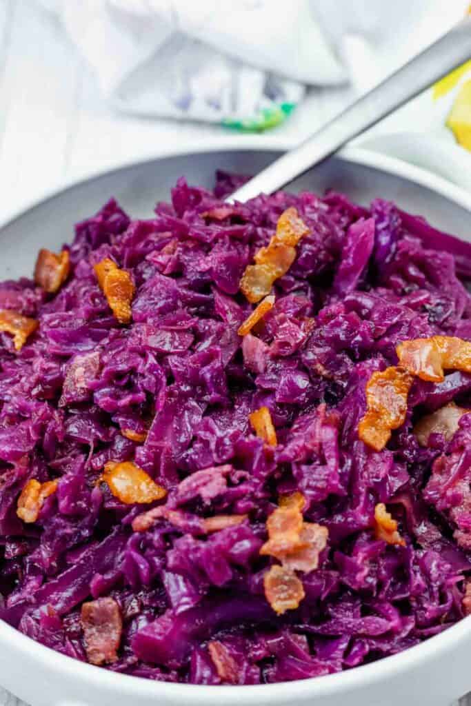 A bowl of cooked red cabbage mixed with pieces of bacon, with a spoon resting in the bowl.