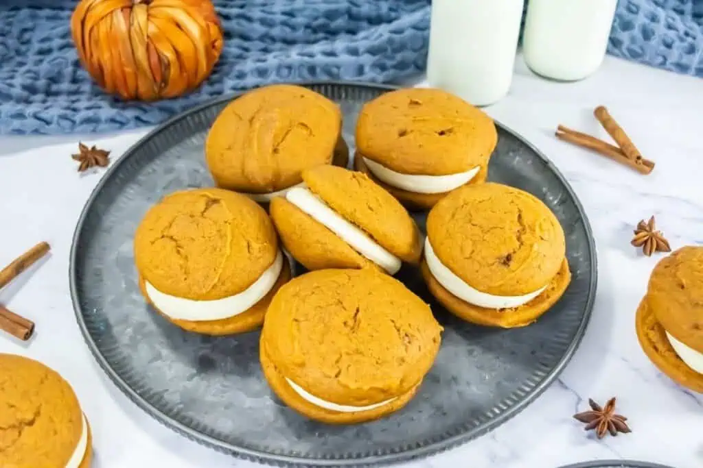 Pumpkin whoopie pies with cream filling on a gray tray with fall spices nearby.