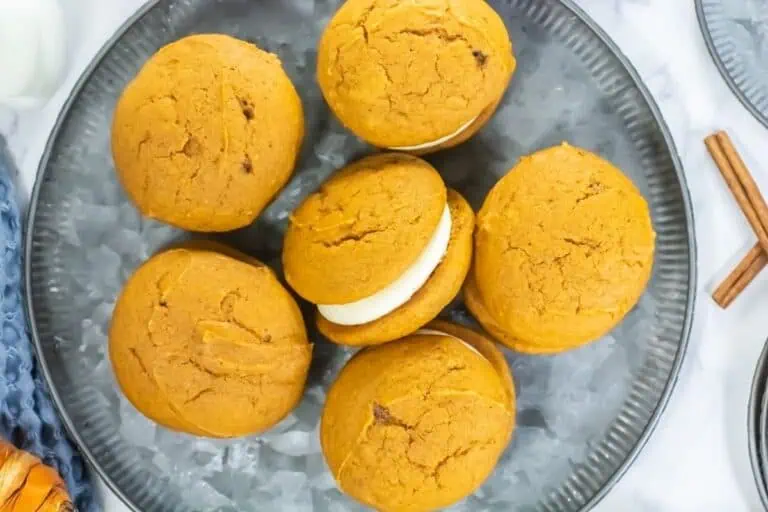 Six pumpkin whoopie pies with cream filling are arranged in a circle on a gray plate, with two cinnamon sticks placed nearby.