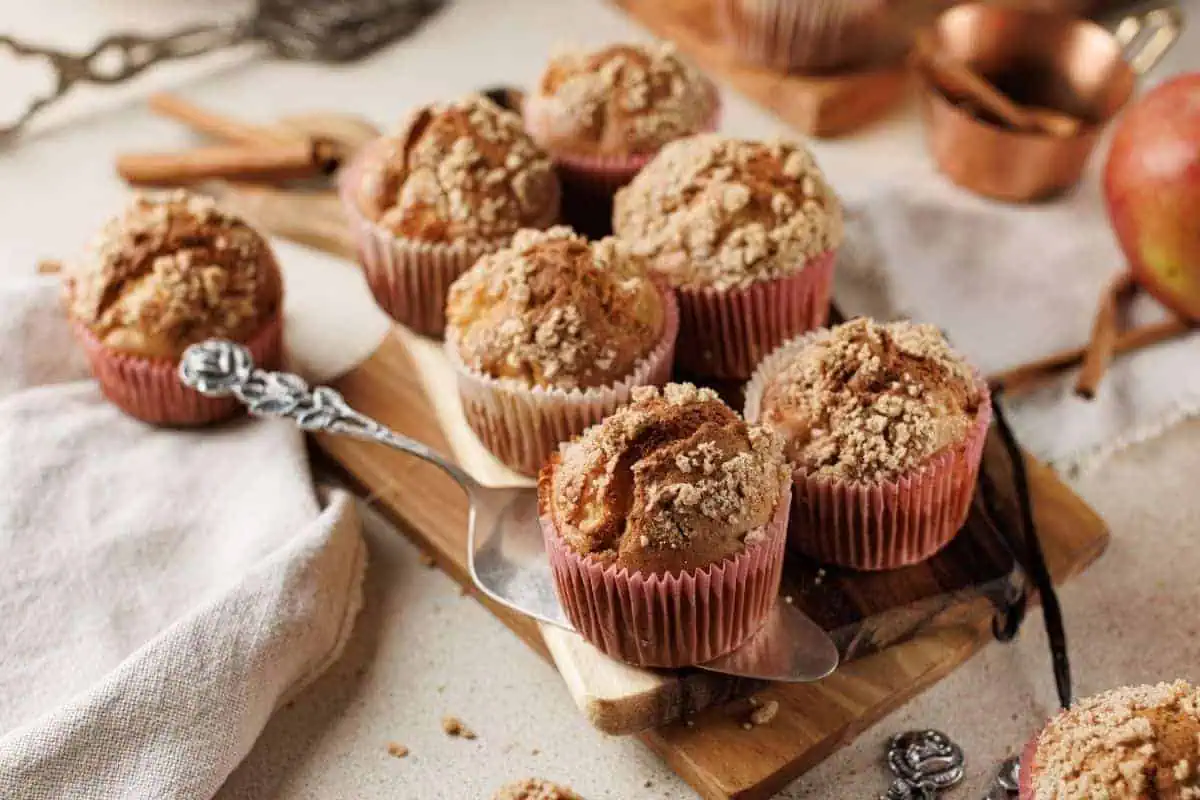 Six Apple Cinnamon Crumb Muffins sit on a wooden board and serving utensil, with baking tools and an apple in the background.