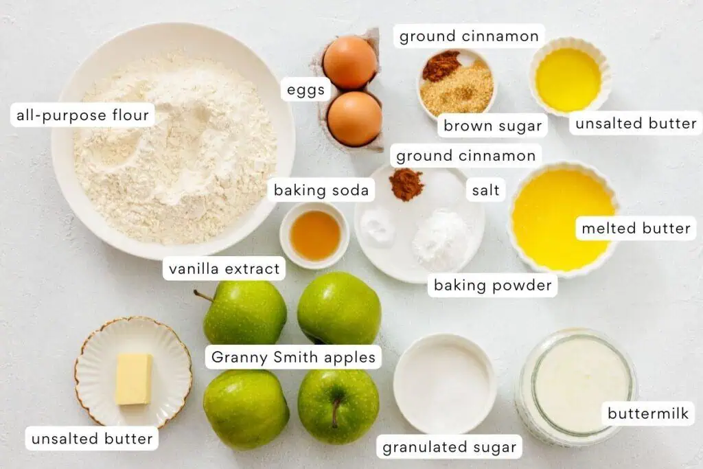 Various labeled baking ingredients arranged on a white surface, including flour, eggs, sugar, butter, apples, spices, buttermilk, and leavening agents.