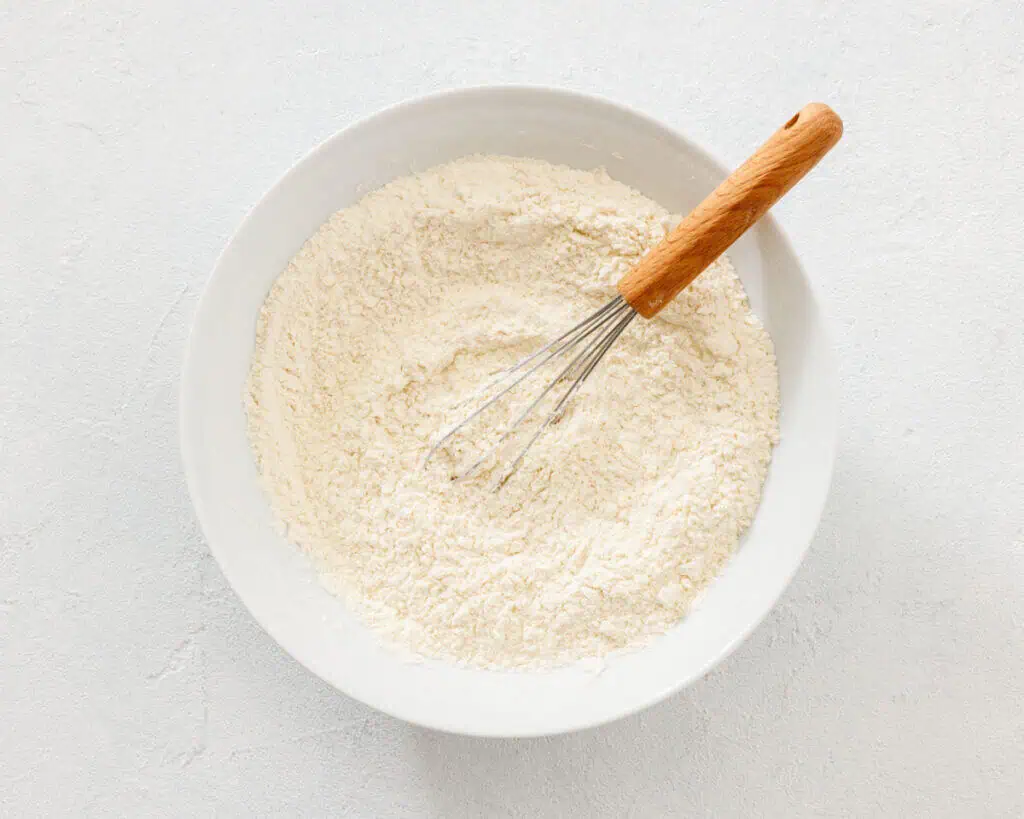 A white bowl filled with flour and a metal whisk with a wooden handle rests inside the bowl on a light surface.