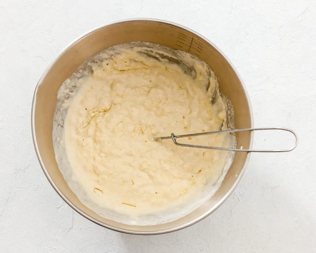 A metal mixing bowl containing a partially mixed batter and a wire whisk, placed on a light-colored surface.