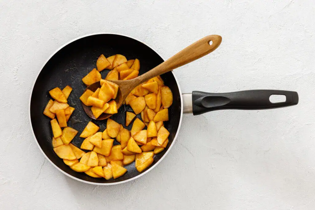 A frying pan with sliced apples being cooked, stirred with a wooden spoon, on a light-colored surface.